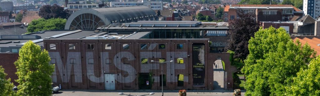 Vue extérieure du musée La Piscine à Roubaix : une façade en briques rouges avec le mot “MUSÉE” peint en grandes lettres, et à l’arrière, l’ancienne verrière de la piscine transformée en espace d’exposition.