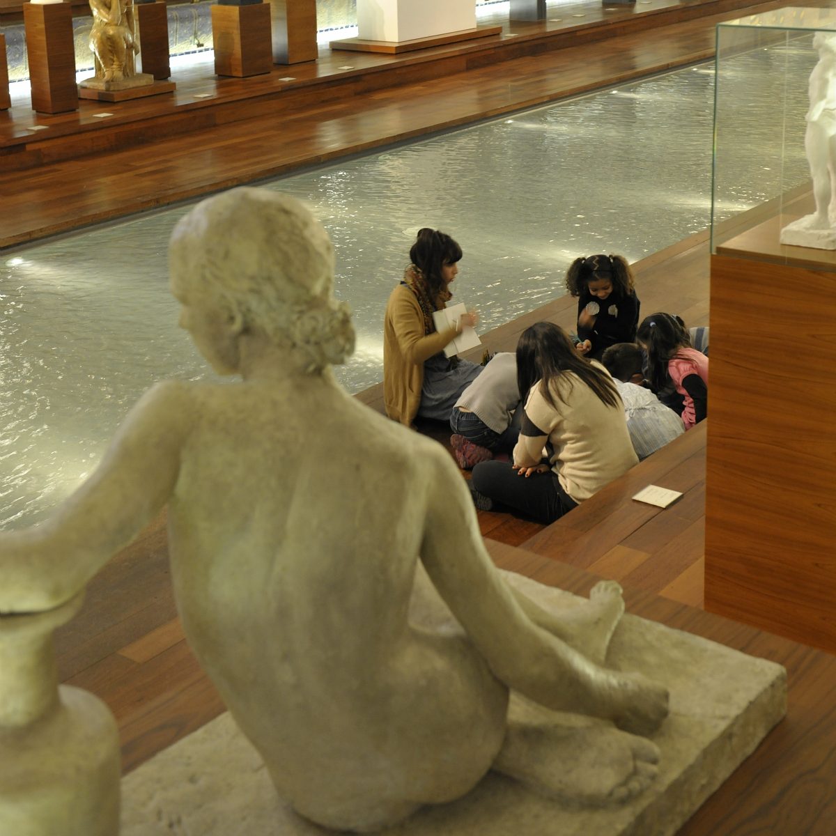 Au bord du bassin du musée La Piscine de Roubaix, une médiatrice culturelle anime un atelier avec des enfants assis au bord de l’ancien bassin, entourés de sculptures exposées autour de l’eau.