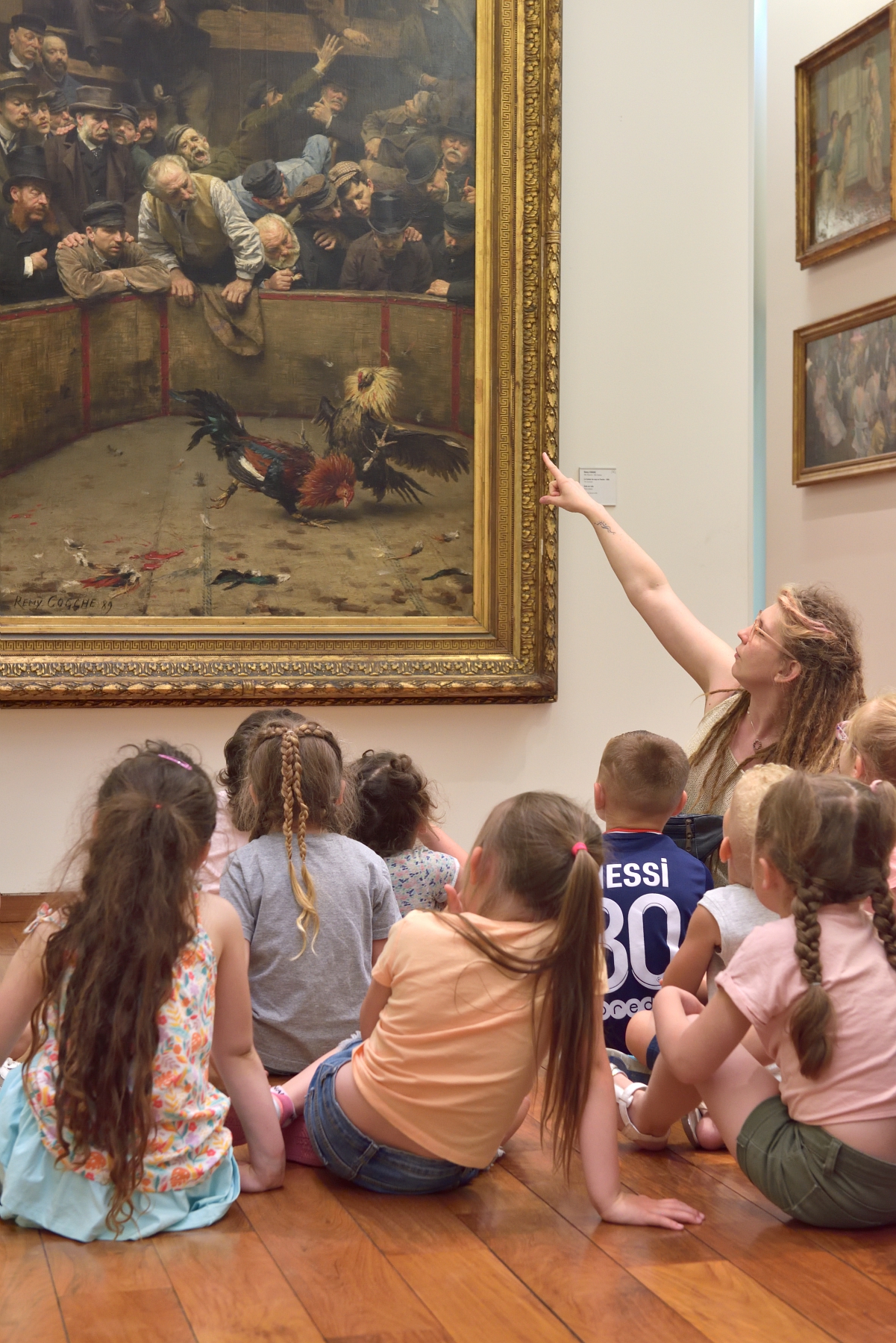Une médiatrice culturelle assise au sol explique le grand tableau représentant le Combat de coqs de Remy Cogghe à un groupe d’enfants attentifs.